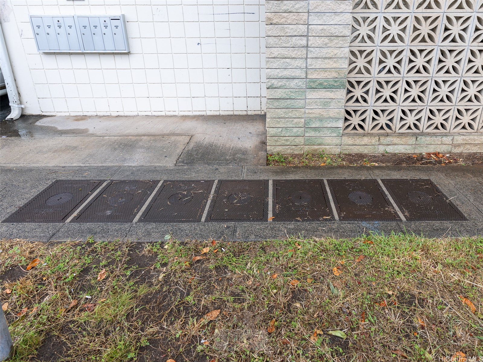 Building exterior with wall-mounted mailboxes, patterned breeze-block wall, and utility covers along the sidewalk.