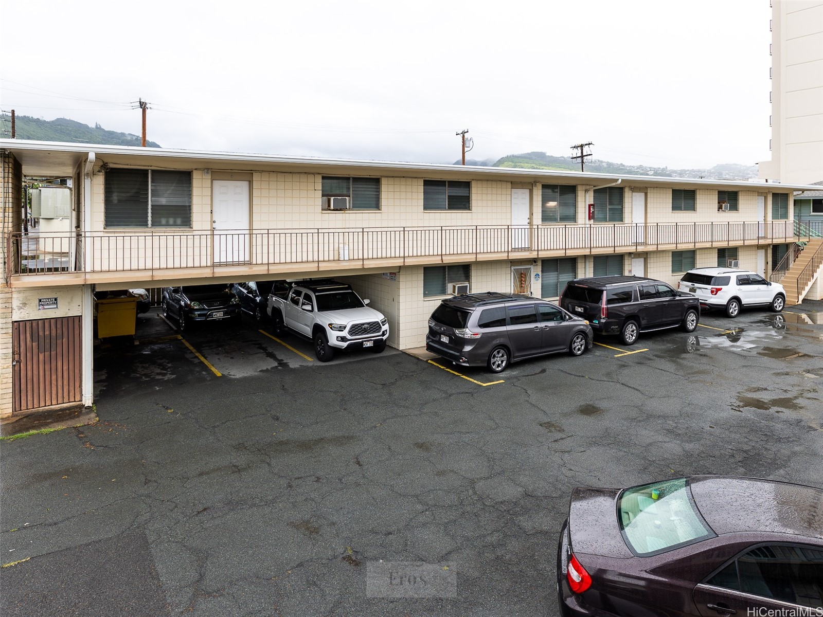 Two-story apartment building with an open-air walkway and parking beneath, backed by green hills.