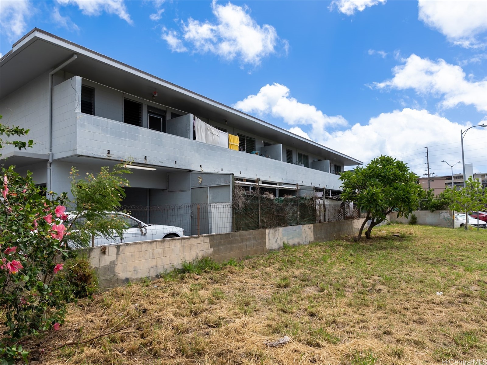 Two-story apartment building with a long row of balconies and a fenced, dry grassy lot.