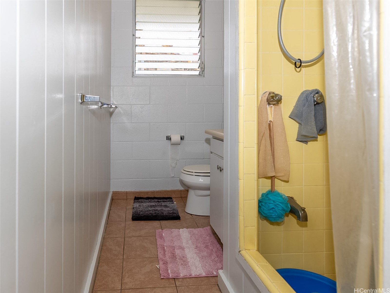 Narrow bathroom with a toilet beneath a louvered window and a yellow-tiled shower.