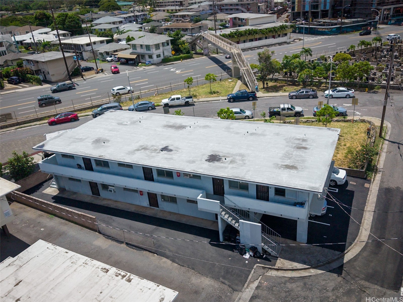 Two-story blue apartment building with a broad flat roof beside a Honolulu road and pedestrian overpass.