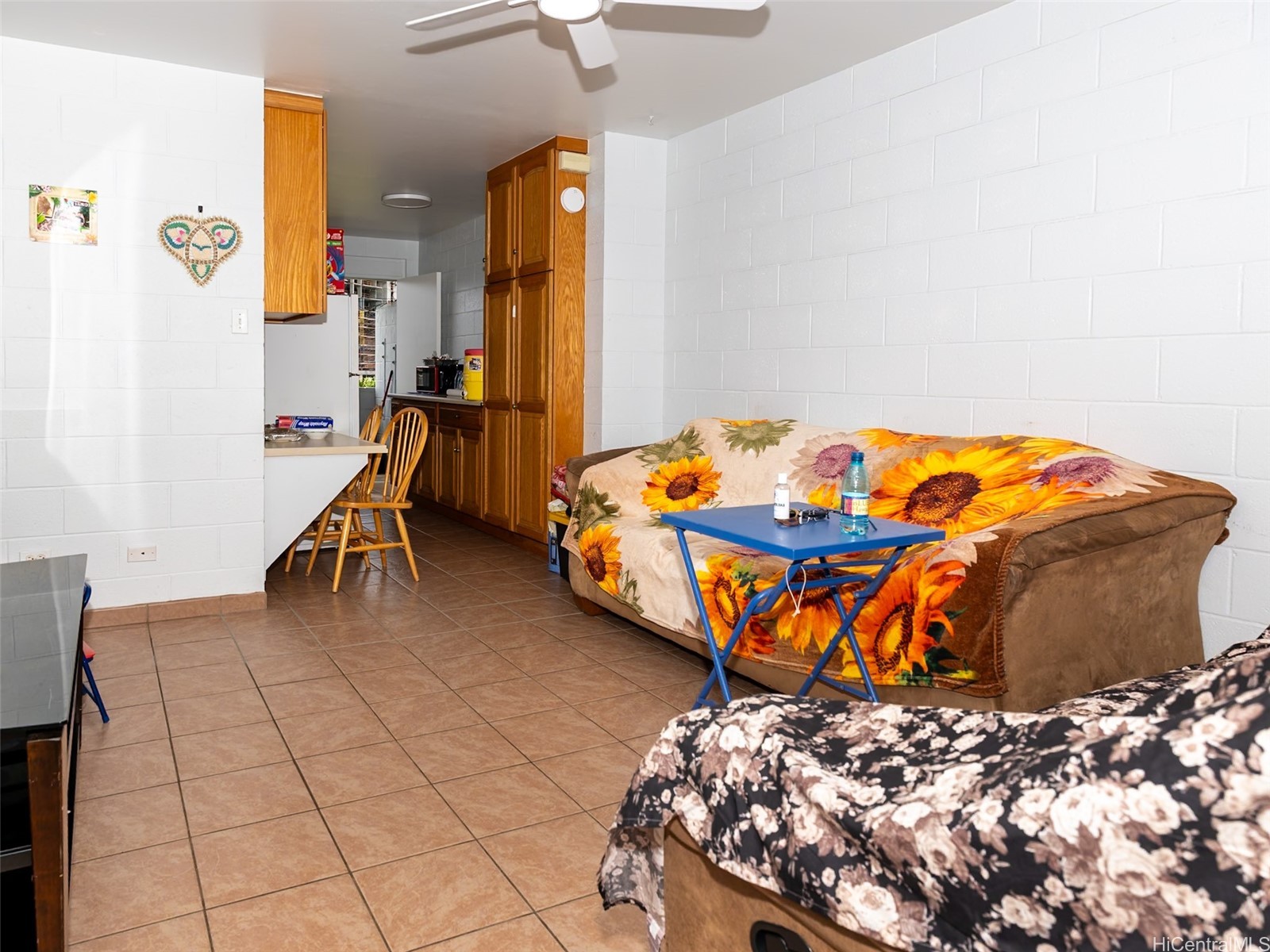 Living area with a sunflower-covered sofa, tiled floor, and compact kitchen with wood cabinets.