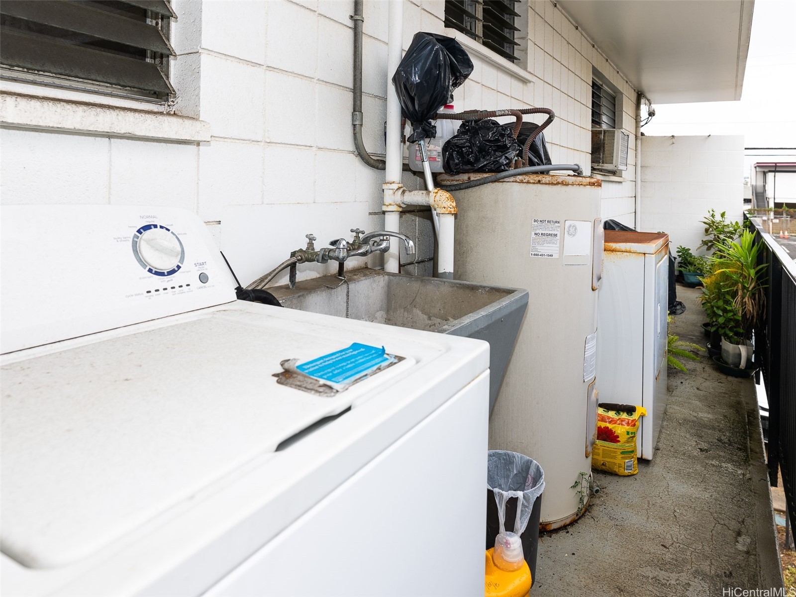 Covered laundry area with washing machines, a utility sink, and potted plants along the walkway.