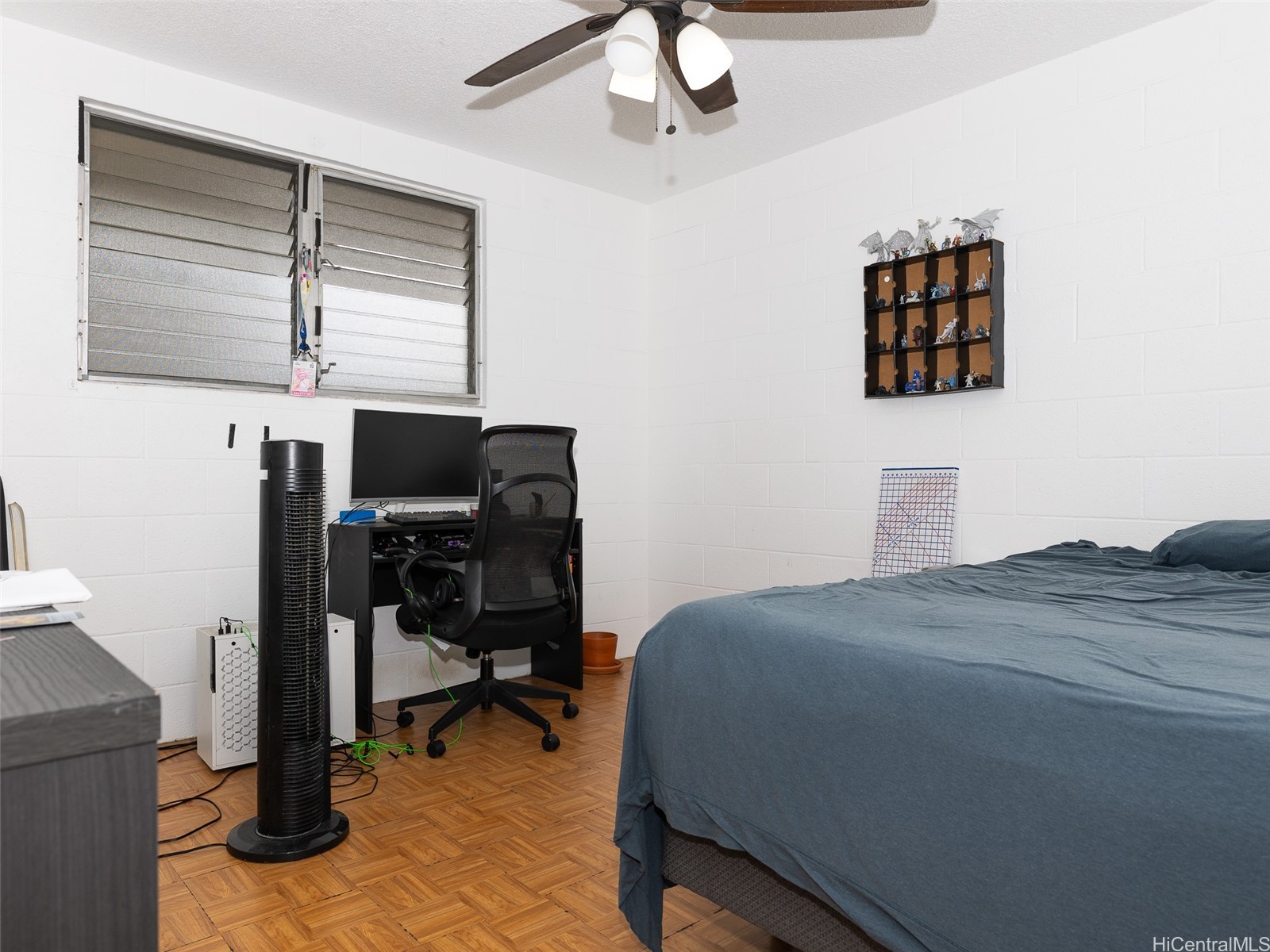 Bedroom with a blue-gray bed, desk setup, louvered windows, and parquet flooring.