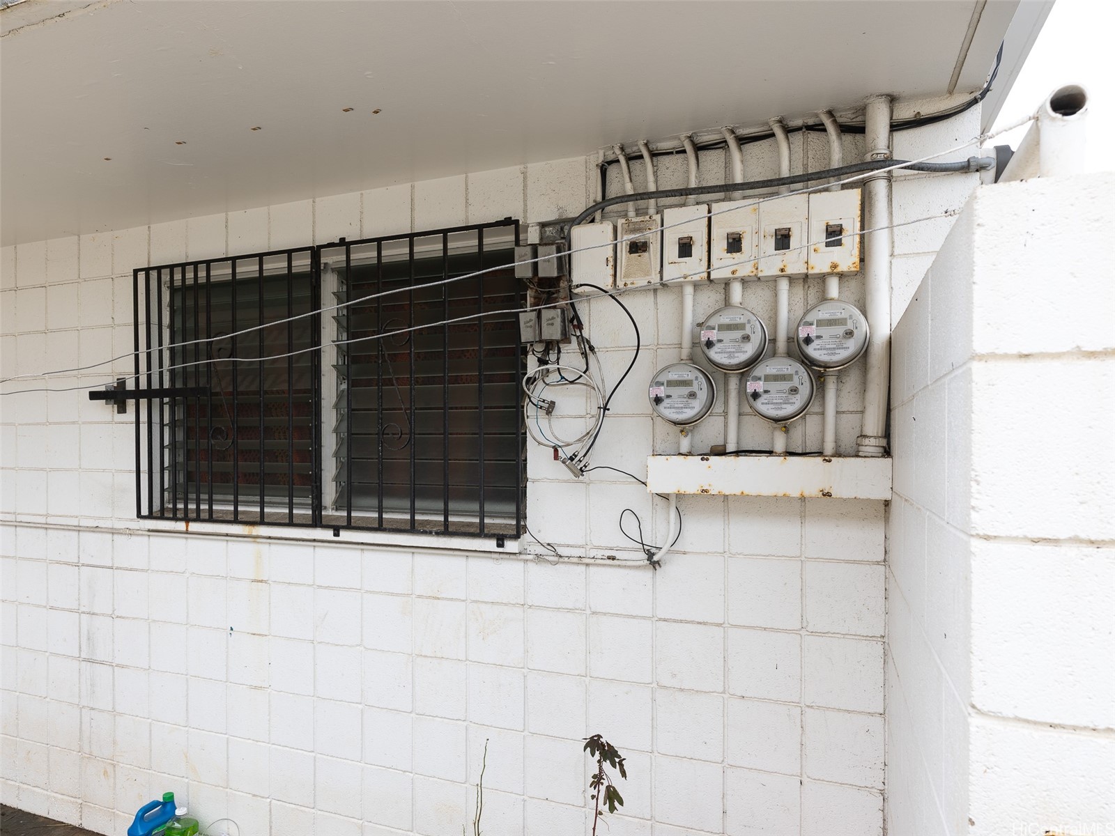 Exterior wall with barred louvered window, electrical meters, panels, and exposed wiring.