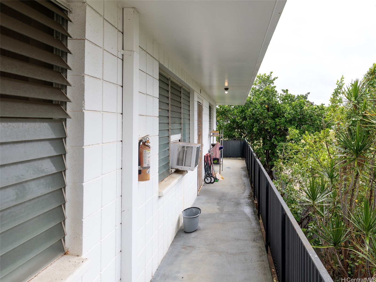 Covered walkway beside a white building, with louvered windows and tropical greenery beyond the railing.