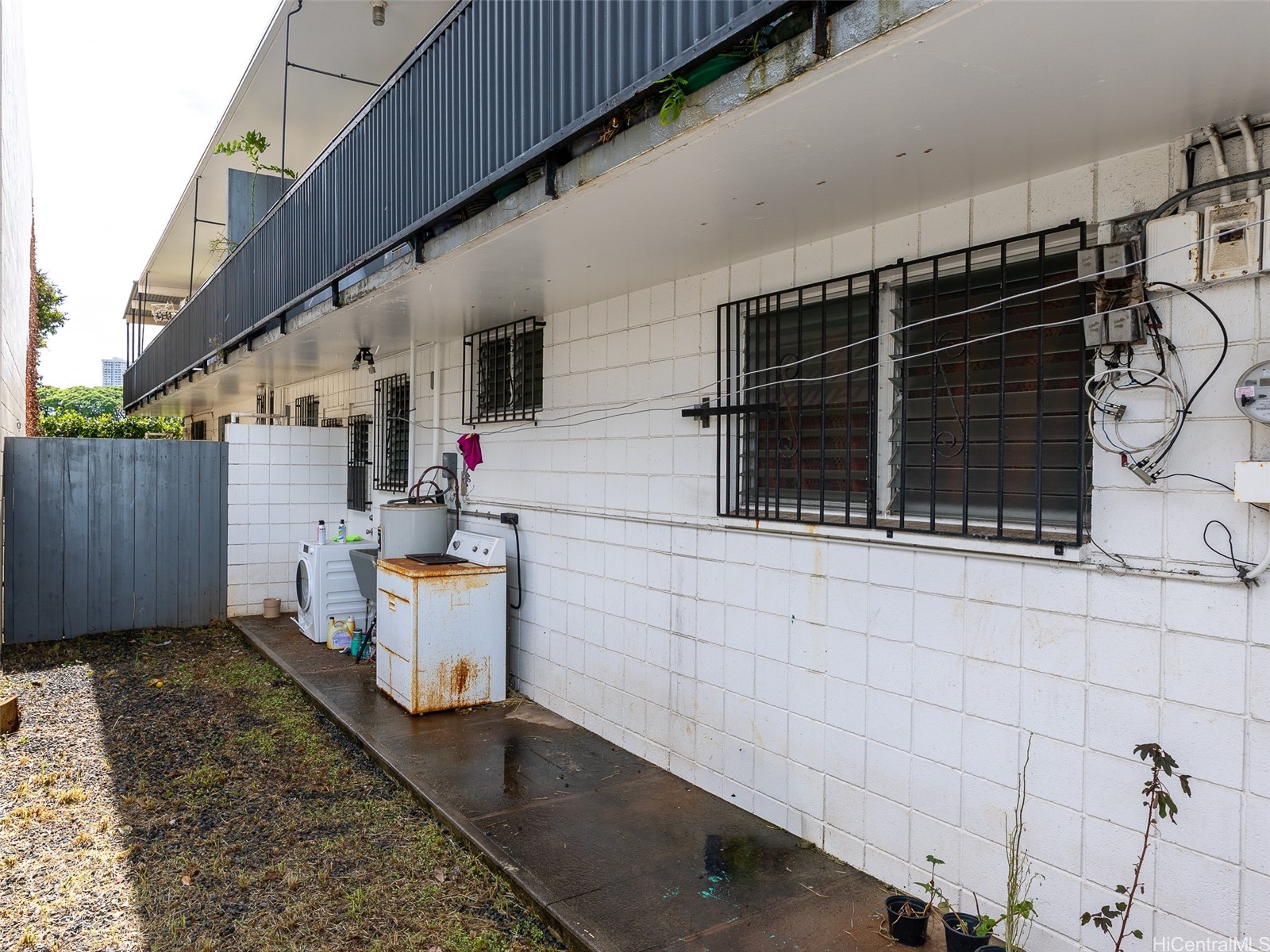 Building exterior with barred windows, an upper balcony, and washing machines beside a paved walkway.