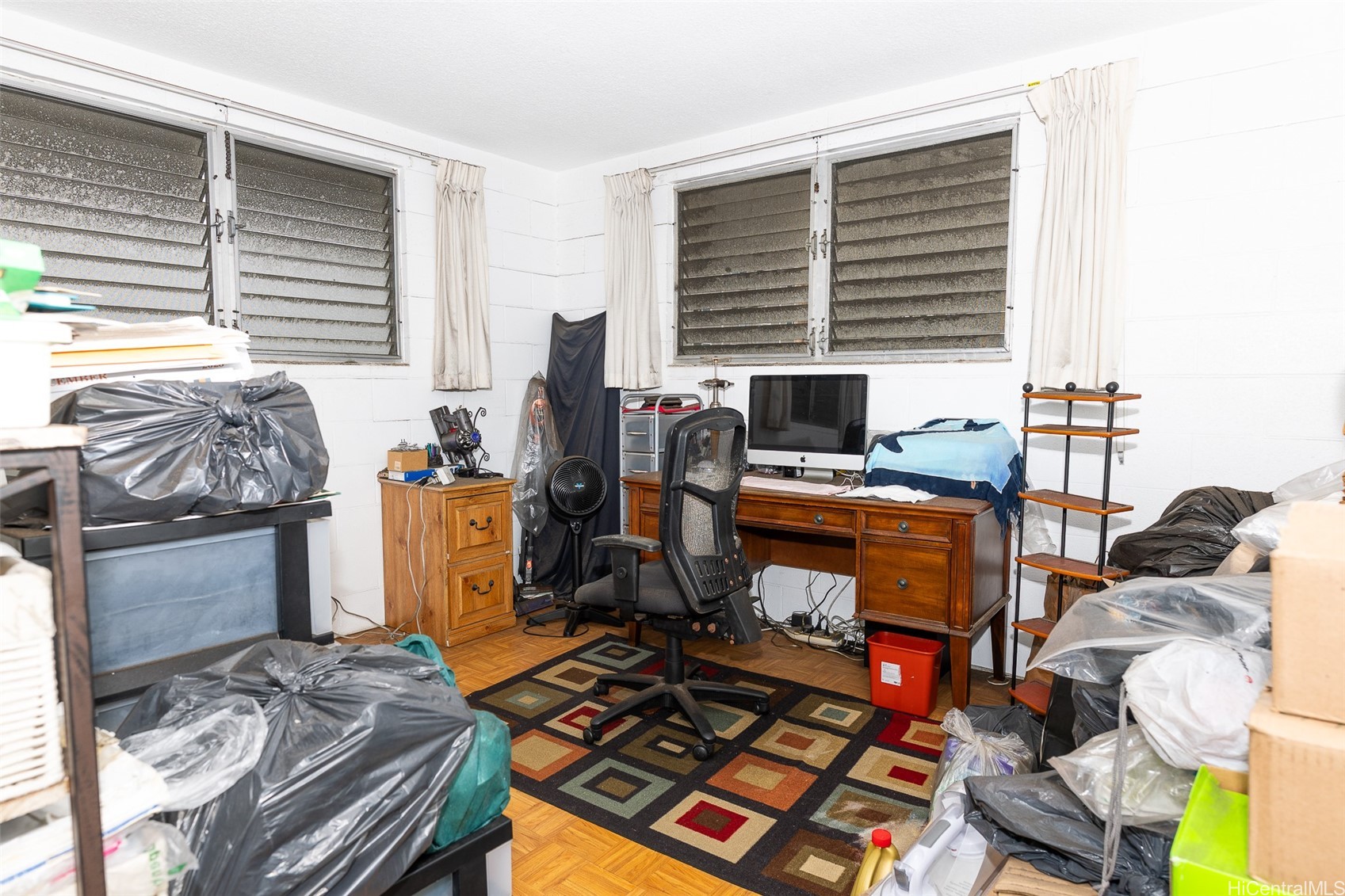 Cluttered office with a wooden desk, computer, louvered windows, and bags and boxes piled around the room.