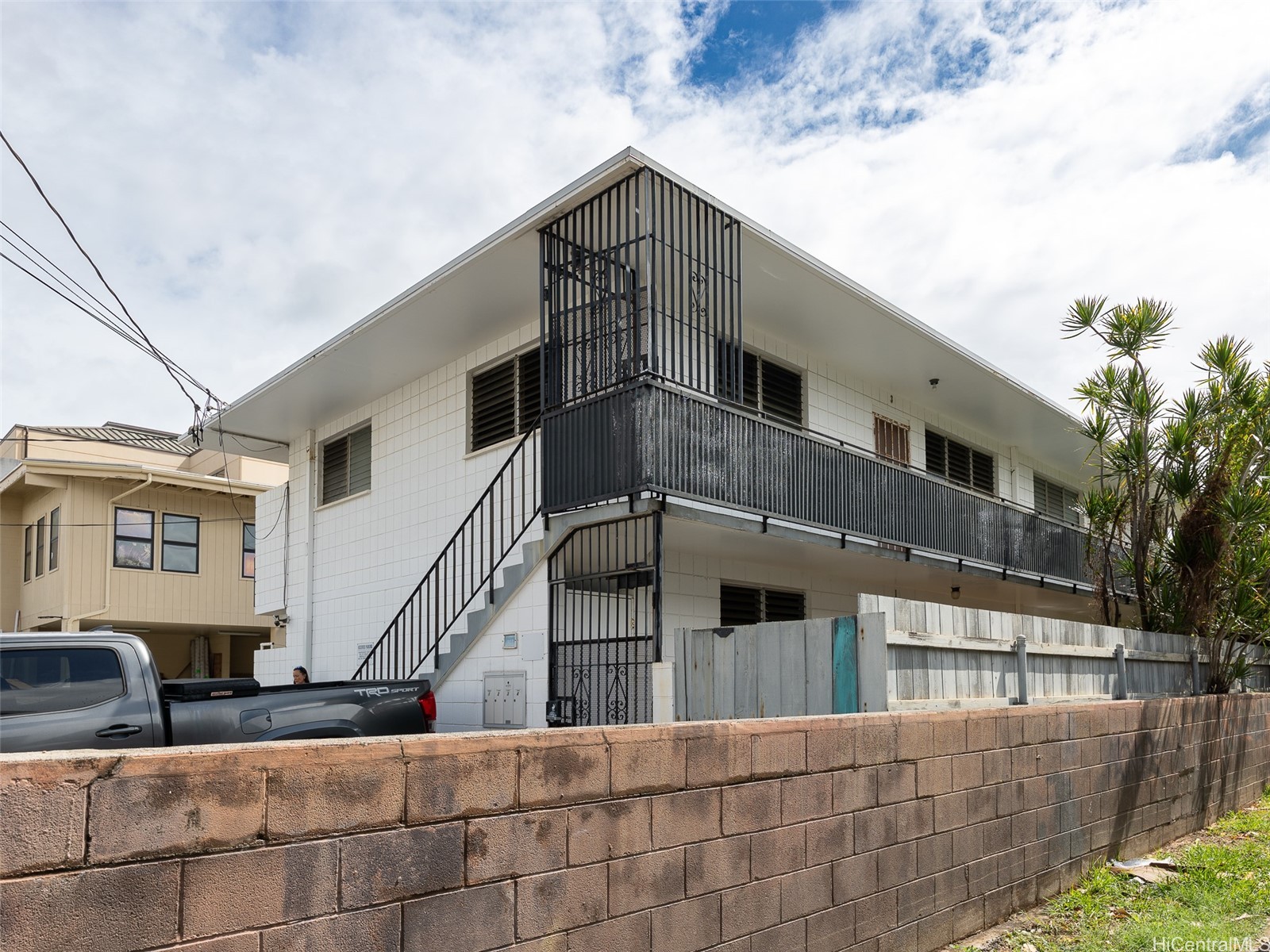 Two-story white apartment building with black metal balconies and an exterior staircase behind a block wall.