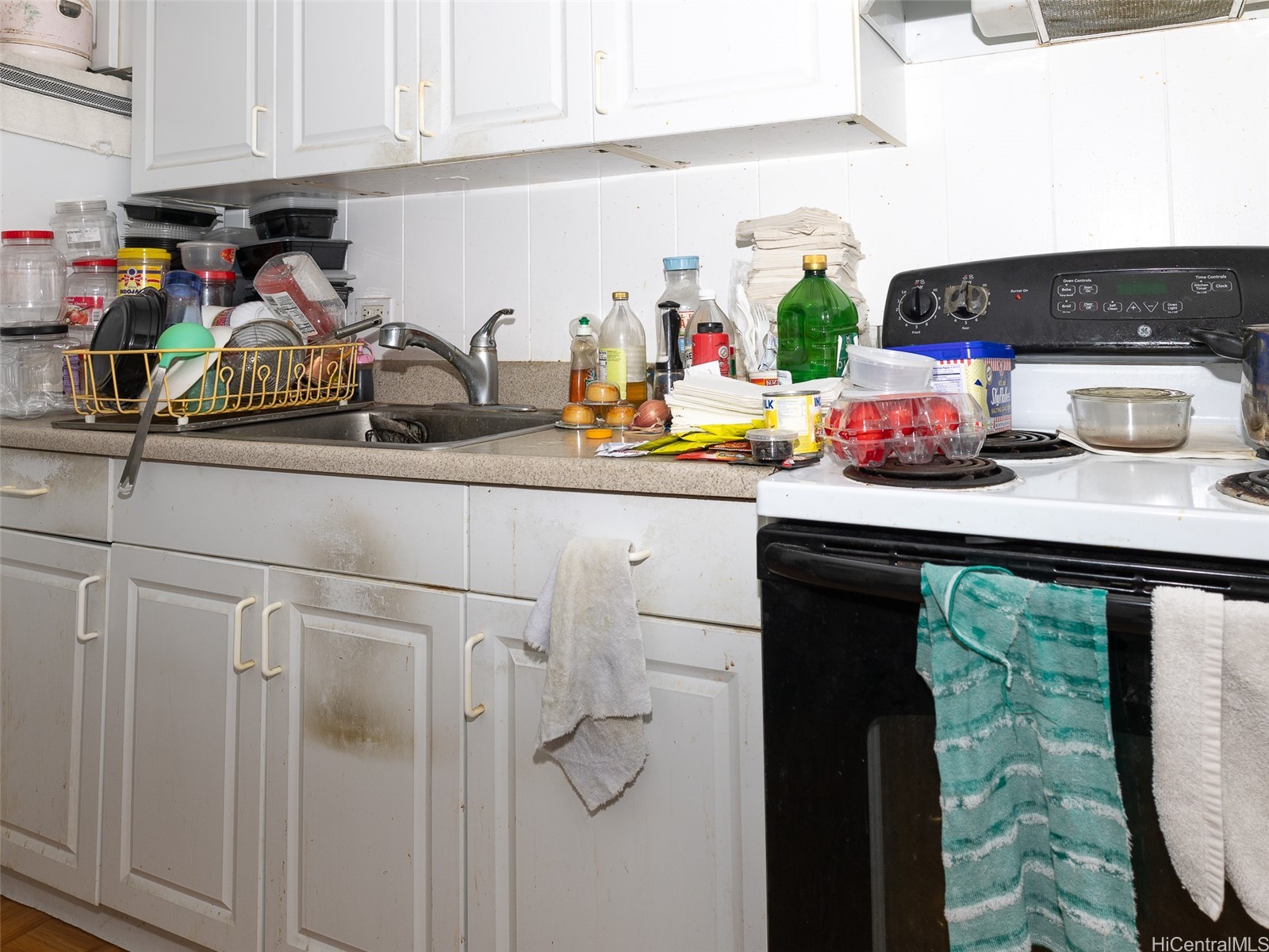 Cluttered kitchen with a dish-filled sink, stained white cabinets, and an electric range with towels.