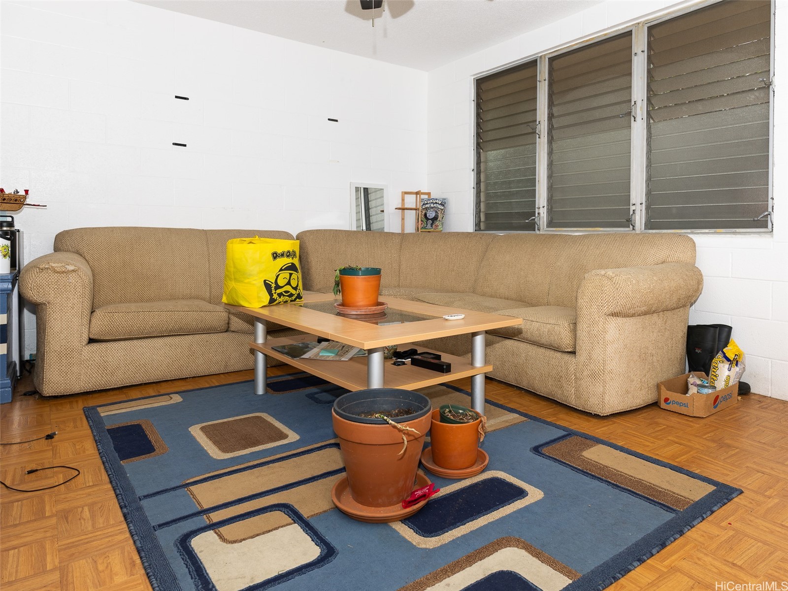 Living room with a tan sectional sofa, coffee table, potted plants, and wide louvered windows.
