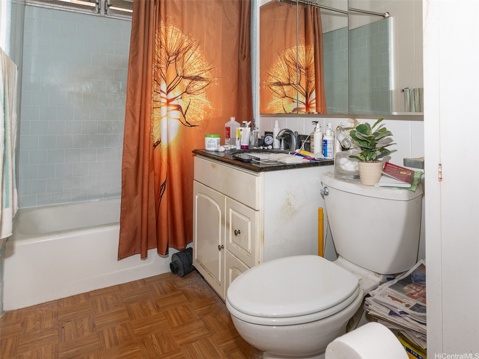 Bathroom with blue-green tile, an orange tree-print shower curtain, mirrored vanity, and parquet flooring.