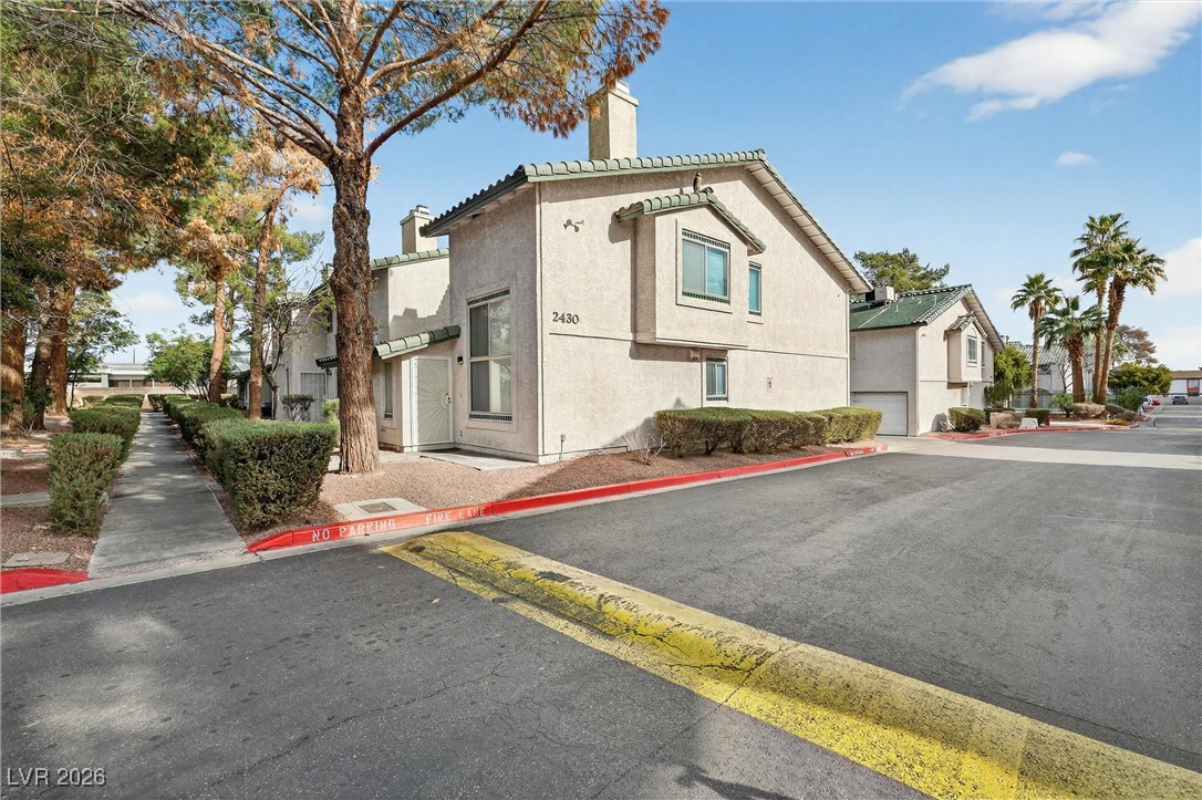 Two-story stucco residence with green tile roofs beside a landscaped sidewalk and red fire-lane curb.