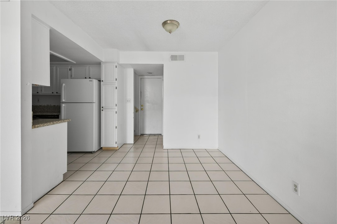 Vacant dining area with square tile floors and an adjoining kitchen with white cabinets and a granite counter.