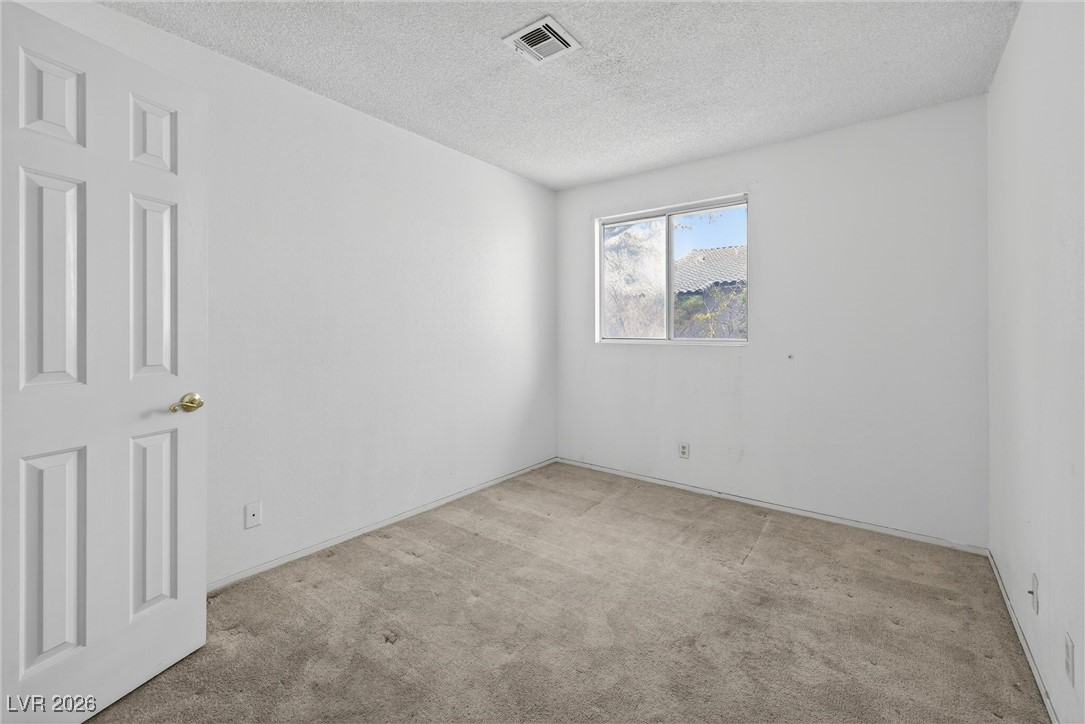 Empty bedroom with beige carpet, white walls, and a window overlooking a neighboring roof.