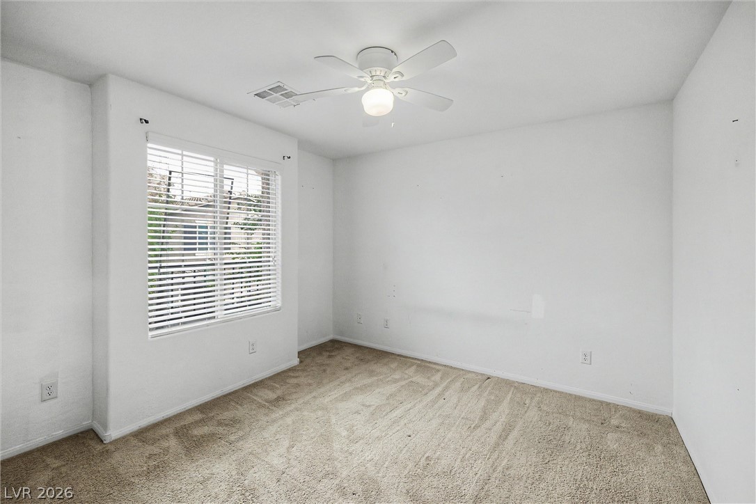 Empty bedroom with beige carpet, a wide window with blinds, and a ceiling fan.