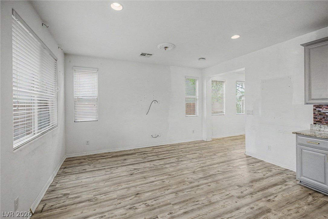 Vacant open room with wood-look flooring, multiple windows, and a partial view of the kitchen.