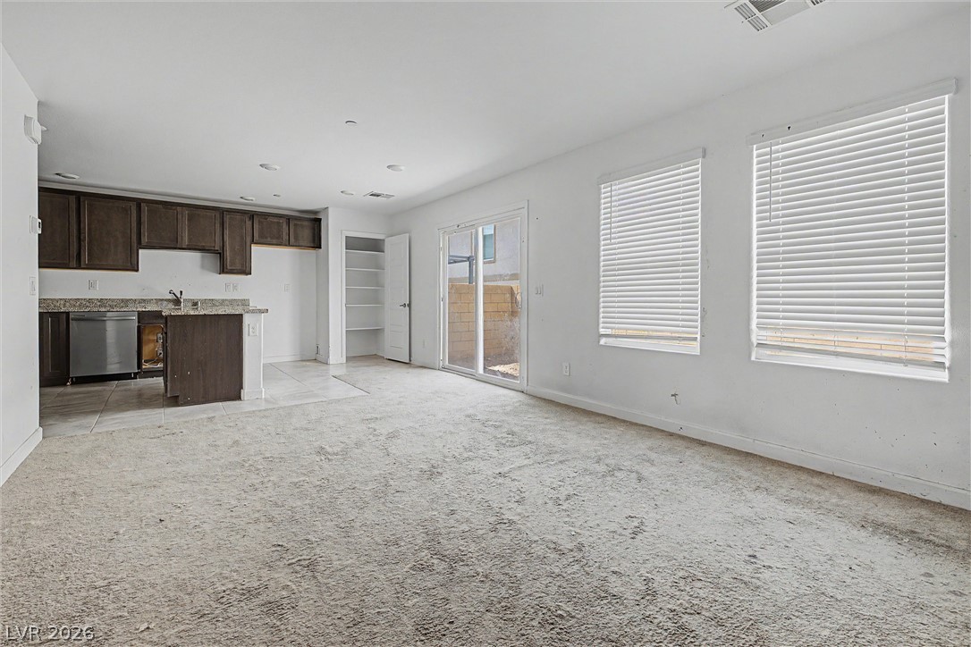 Empty carpeted living room with an open kitchen, two blinded windows, and a sliding glass door.