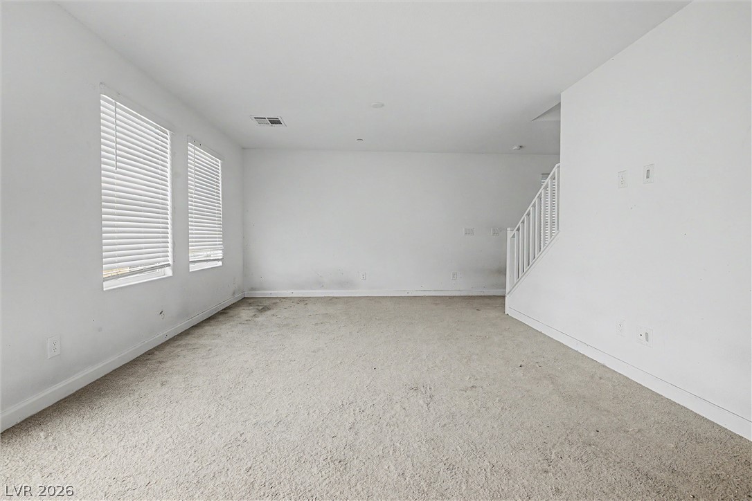 Empty living room with carpet, two windows with blinds, and a staircase.