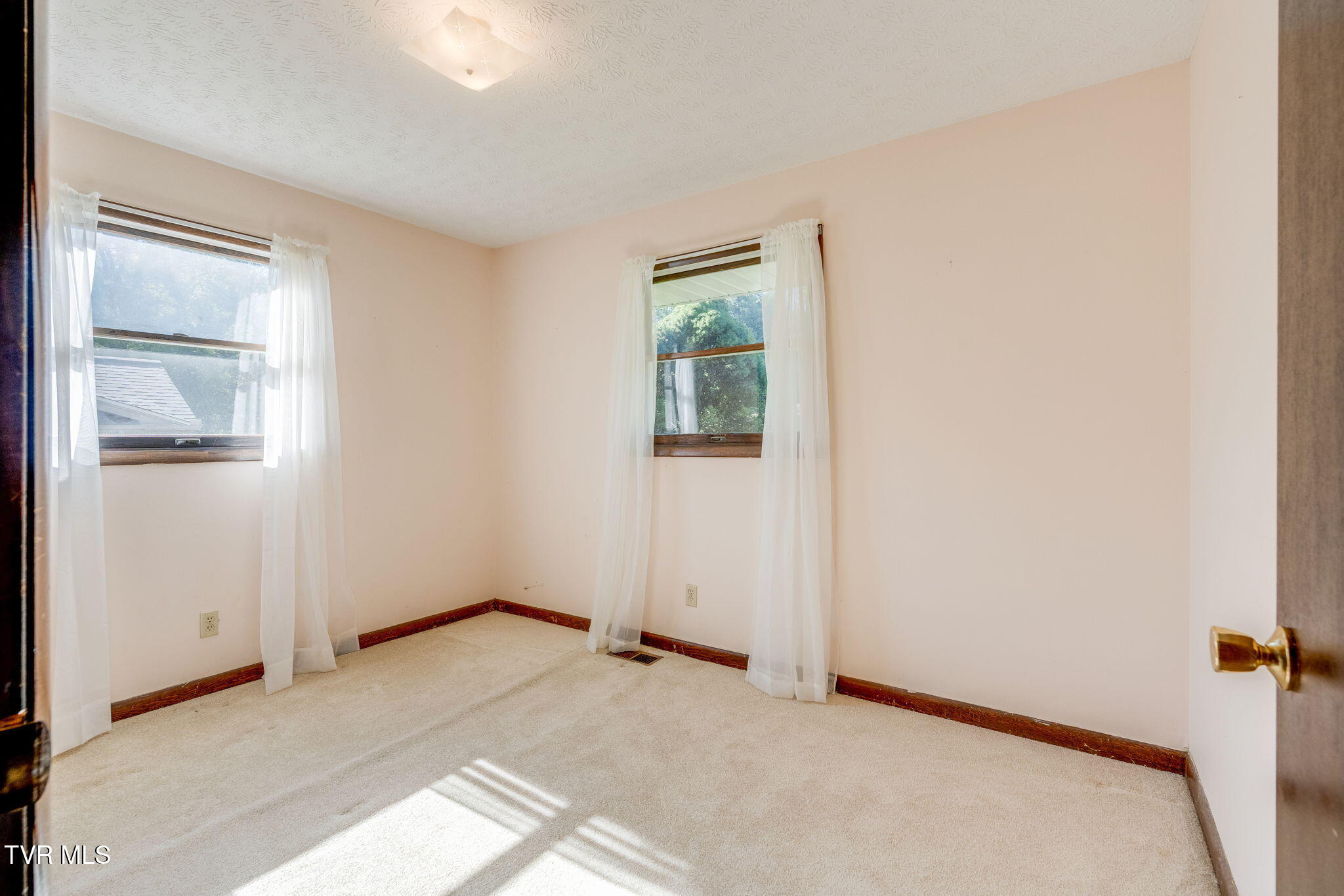 Empty bedroom with beige carpet, two curtained windows, and sunlight across the floor.