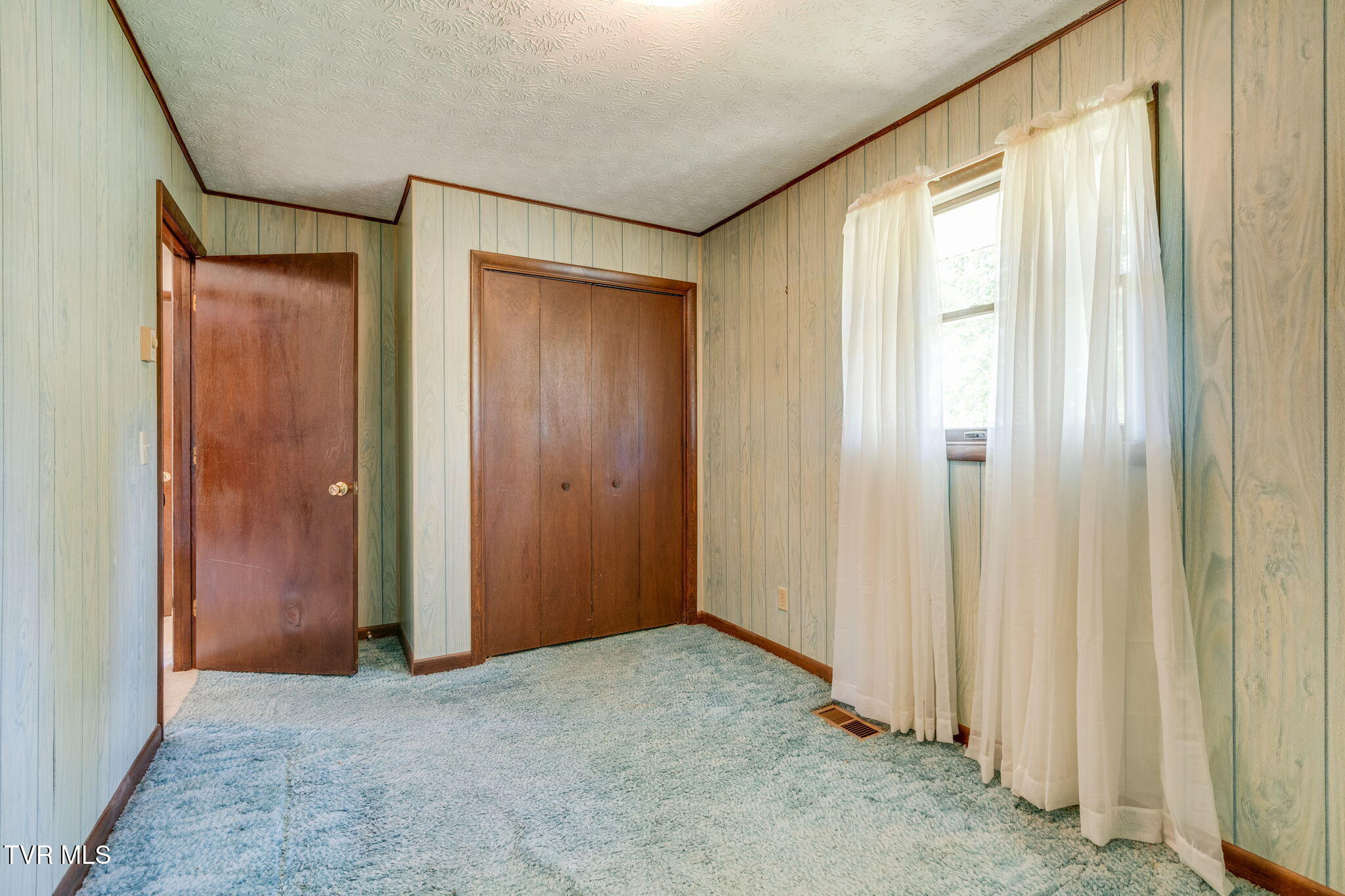 Empty bedroom with a brown double closet, blue-gray carpet, and sheer curtains over the window.