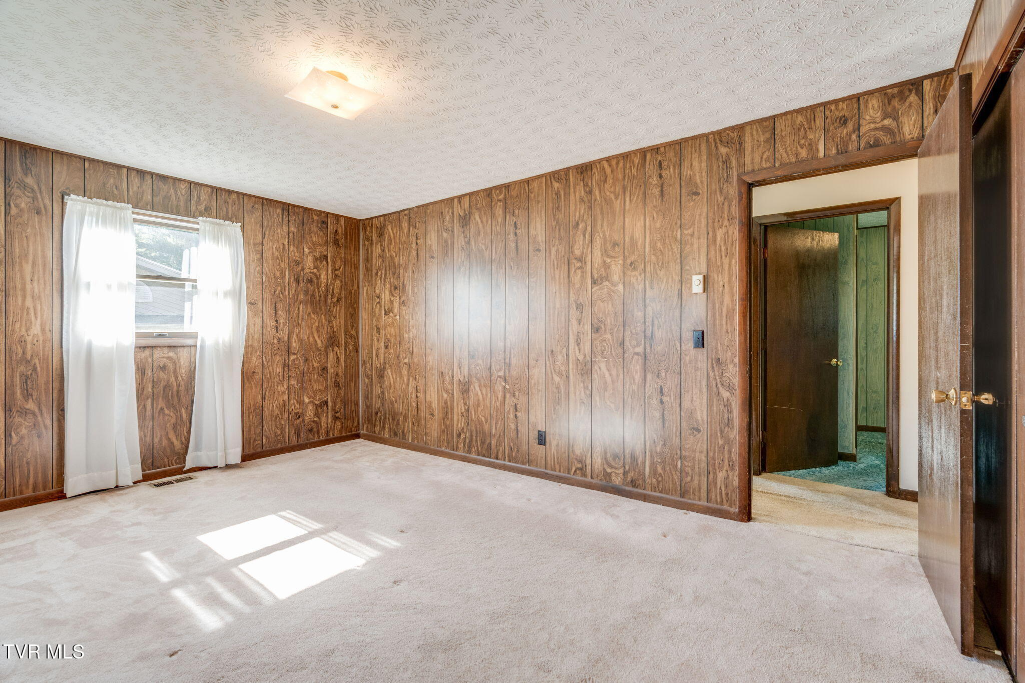 Empty carpeted room with wood-paneled walls, a curtained window, and an open doorway.