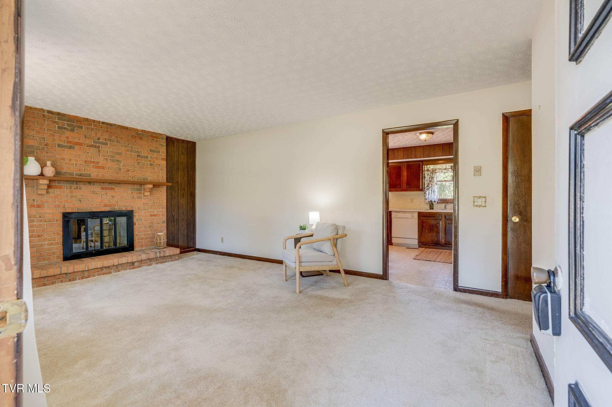 Living room with a brick fireplace, beige carpet, and doorway to the kitchen.