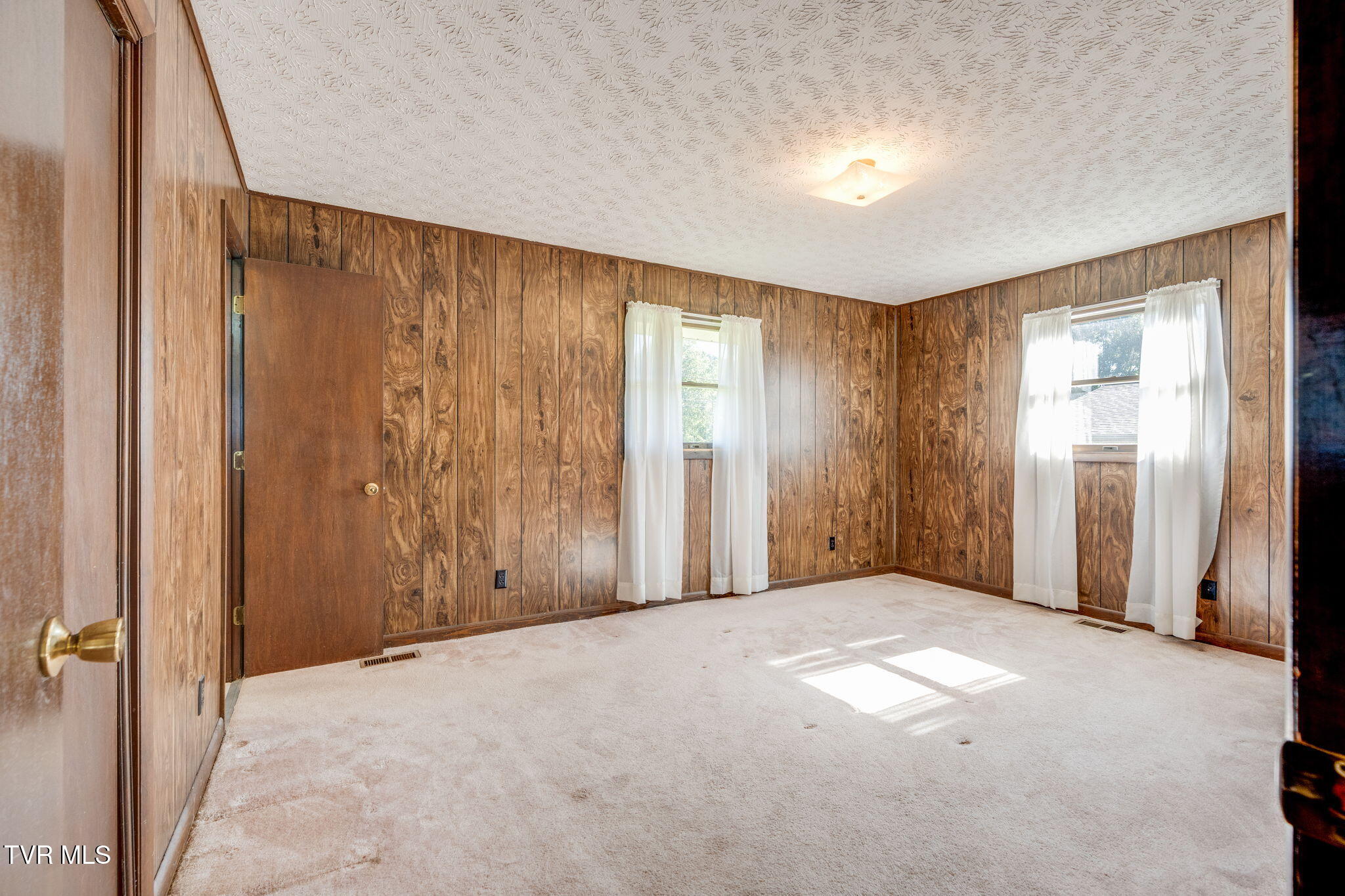 Empty room with wood-paneled walls, light carpet, and two curtained windows.