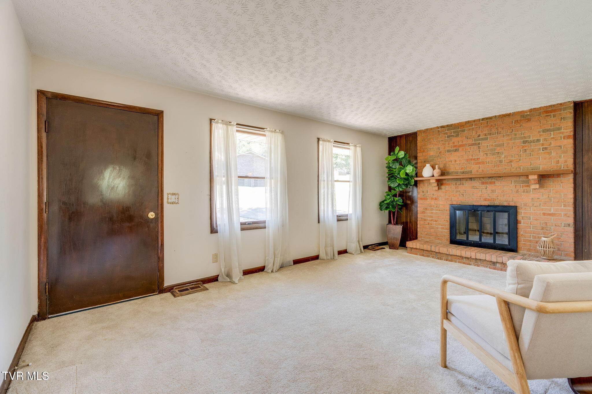 Living room with a brick fireplace, two curtained windows, beige carpet, and a dark wooden door.