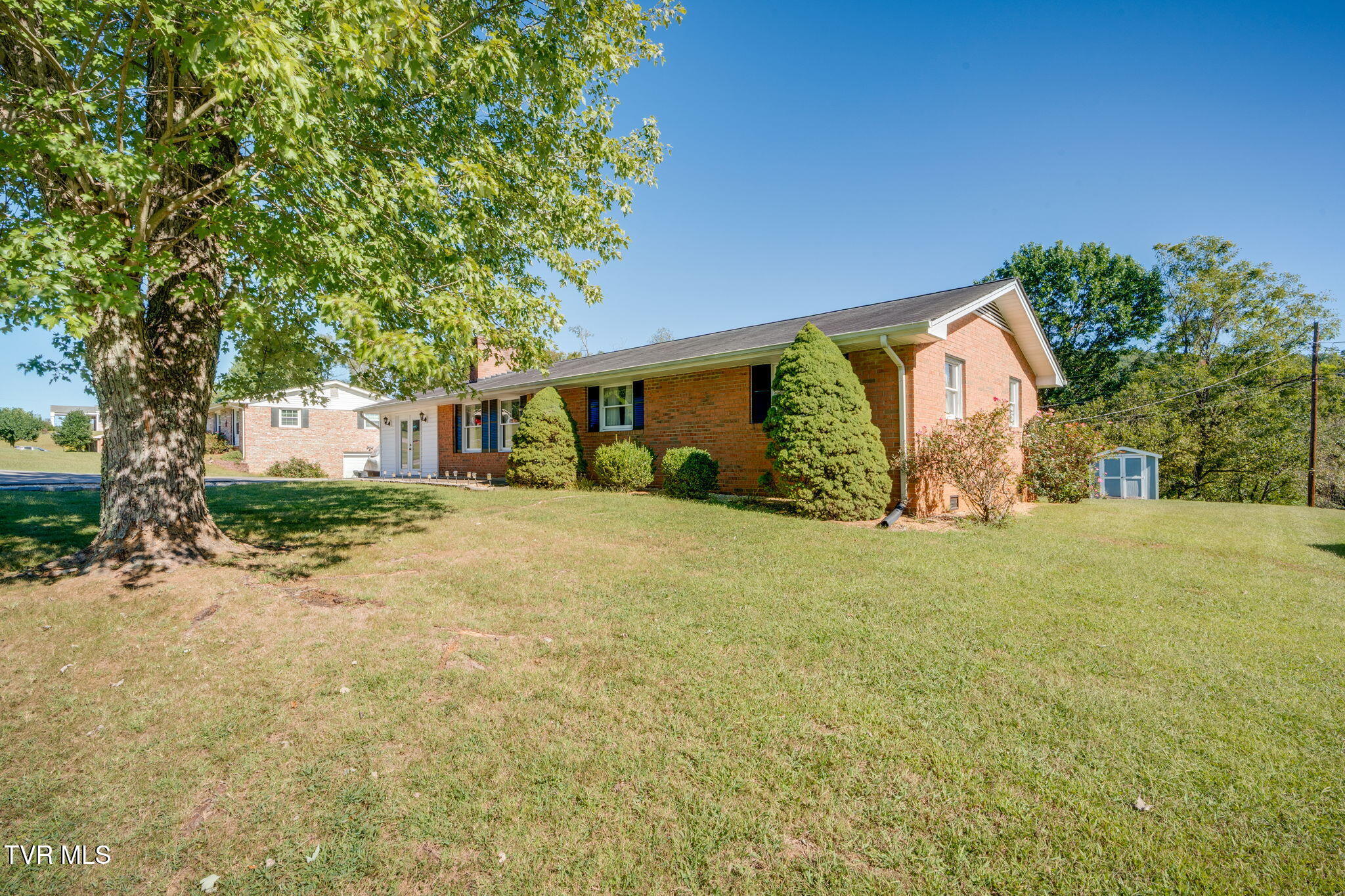 Red brick ranch house with dark shutters, a broad lawn, mature shade tree, and small side-yard shed.
