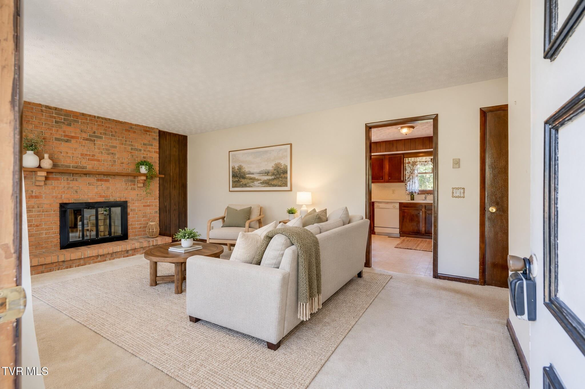 Living room with a brick fireplace, cream sofas, and an adjoining kitchen.