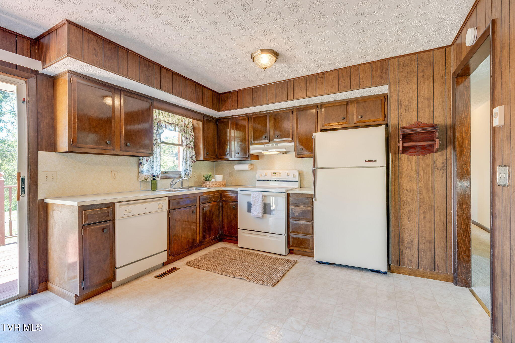 Kitchen with dark wood cabinets, white appliances, and a sink beneath a curtained window.