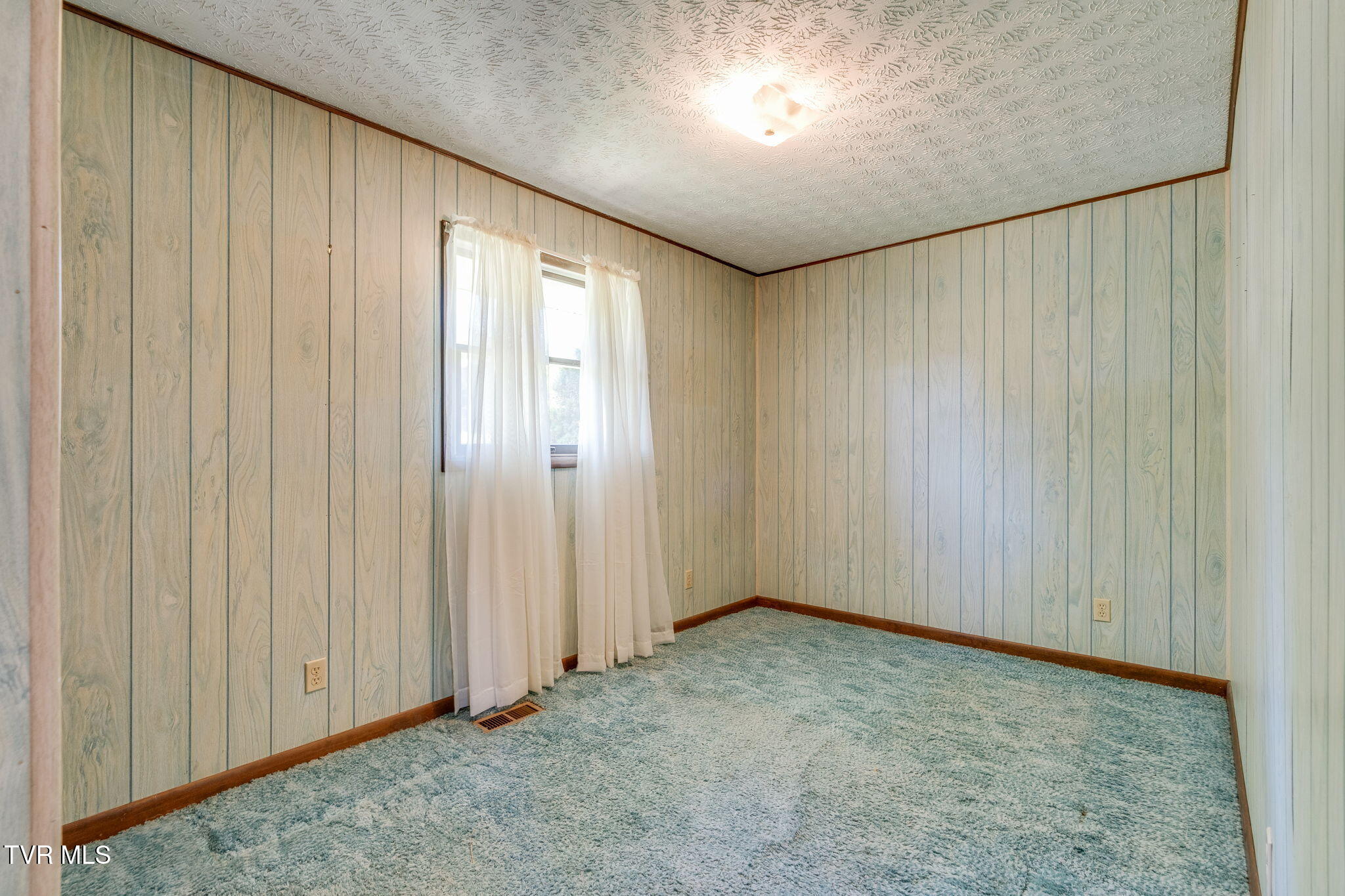 Empty carpeted room with pale paneling and a curtained window.