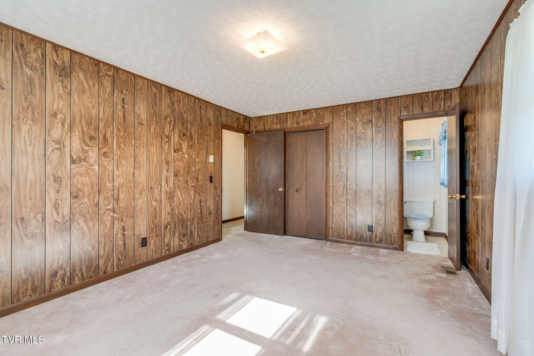 Empty bedroom with wood-paneled walls and carpet, adjoining a bathroom.
