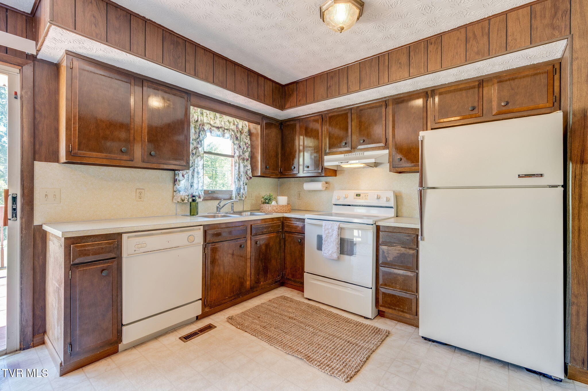 Kitchen with dark wood cabinets, white appliances, and a double sink beneath a curtained window.