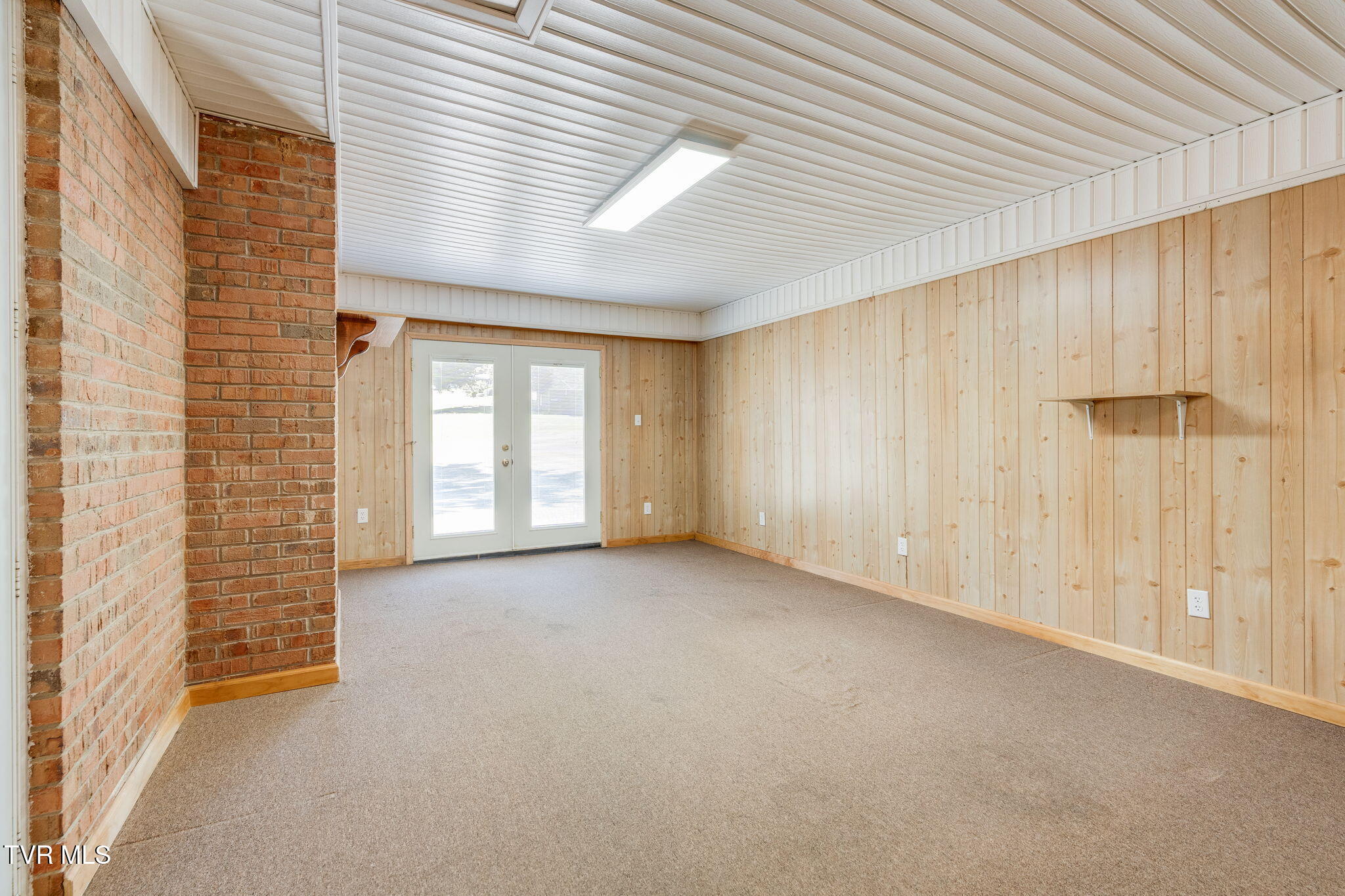 Empty carpeted room with wood-paneled walls, a brick partition, and glazed double doors.