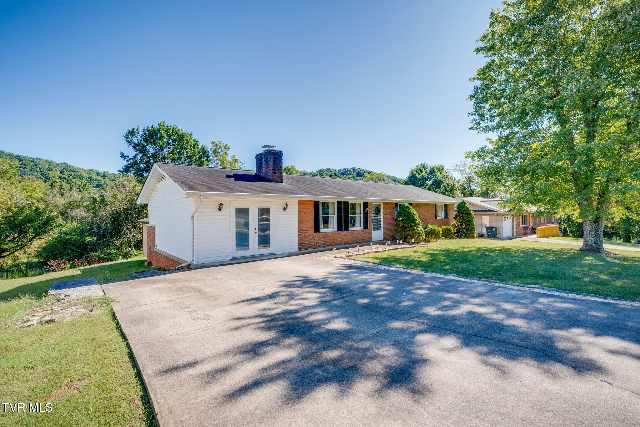 Single-story brick and white-sided home with a broad concrete driveway and wooded hills behind it.