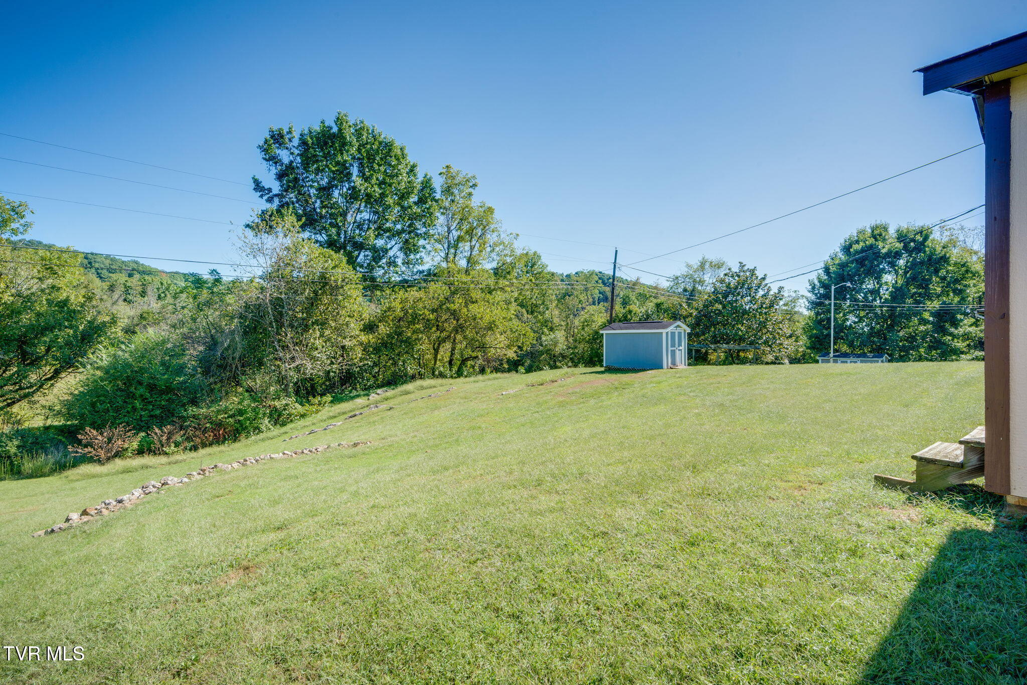Sloping grassy backyard with a blue shed, stone border, and wooded edge.