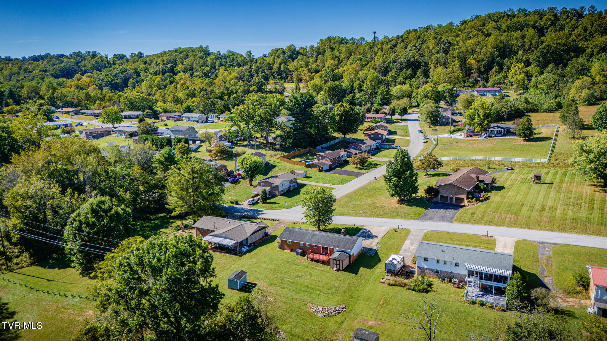 Aerial view of homes along neighborhood streets, with wooded hills and a fenced grassy field.