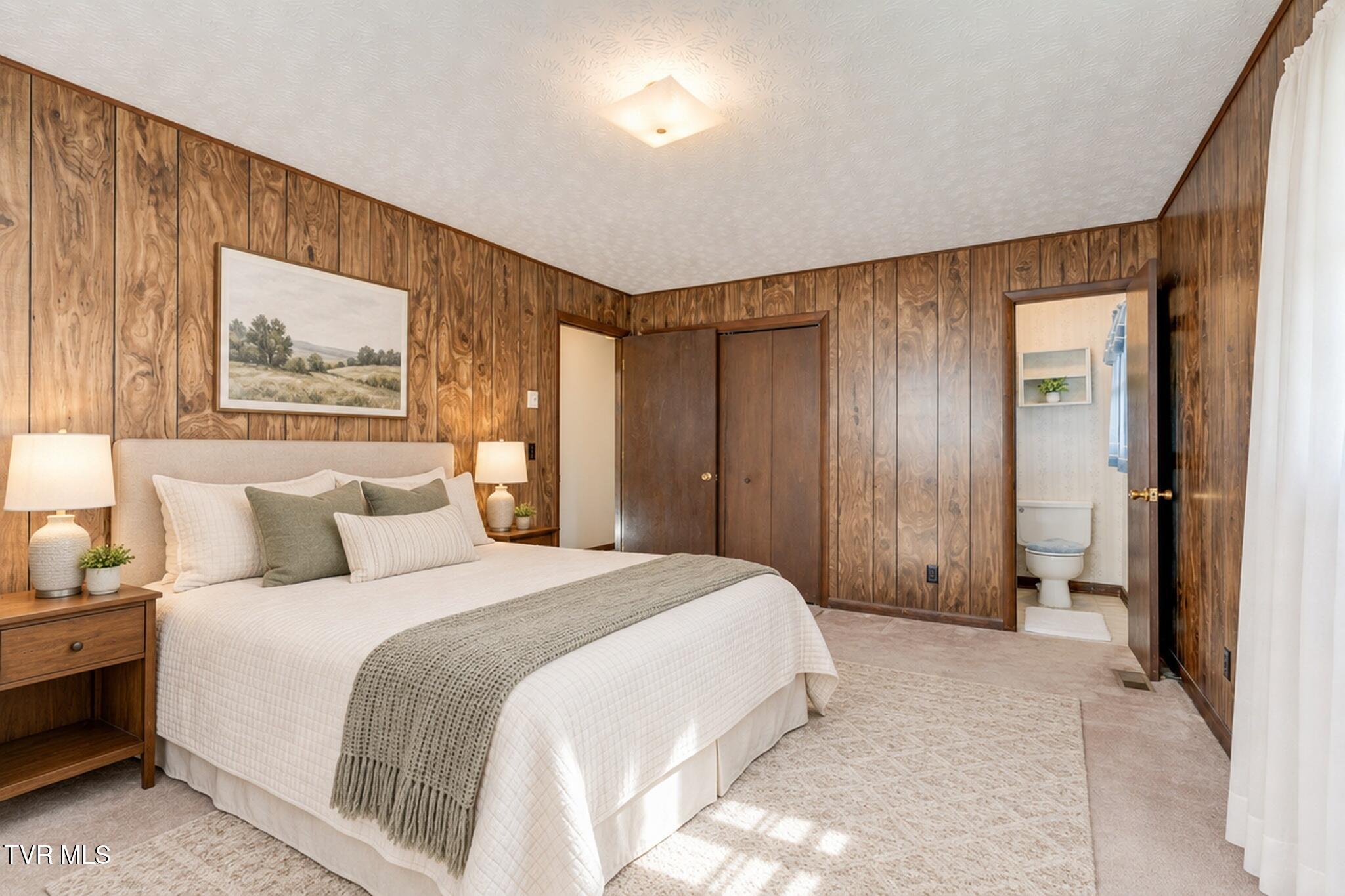 Furnished bedroom with wood-paneled walls, a large white bed, and an open bathroom doorway.