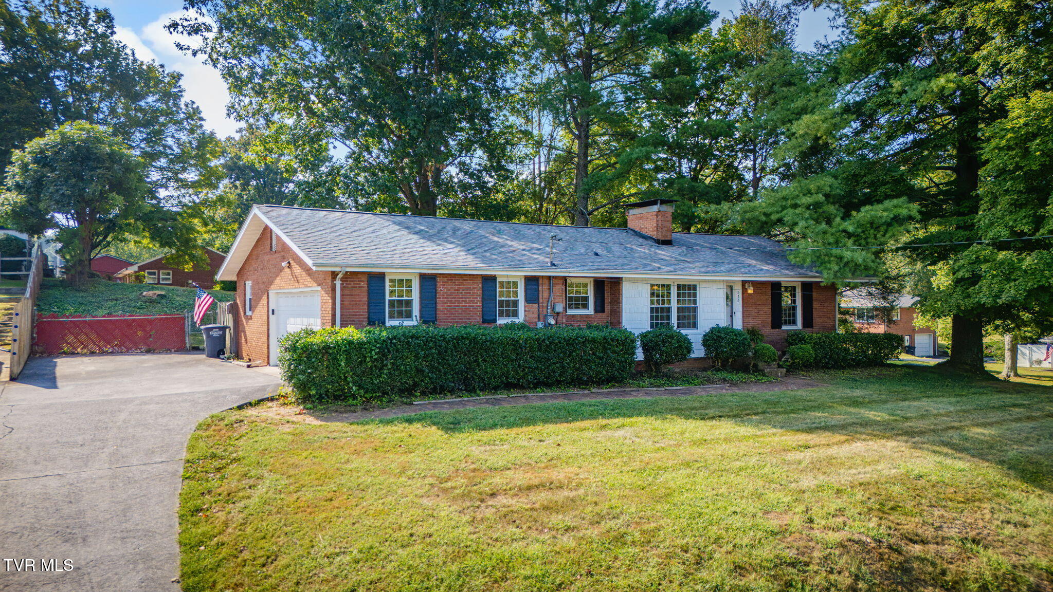 Brick ranch house with an attached garage, broad driveway, front shrubs, and mature shade trees.