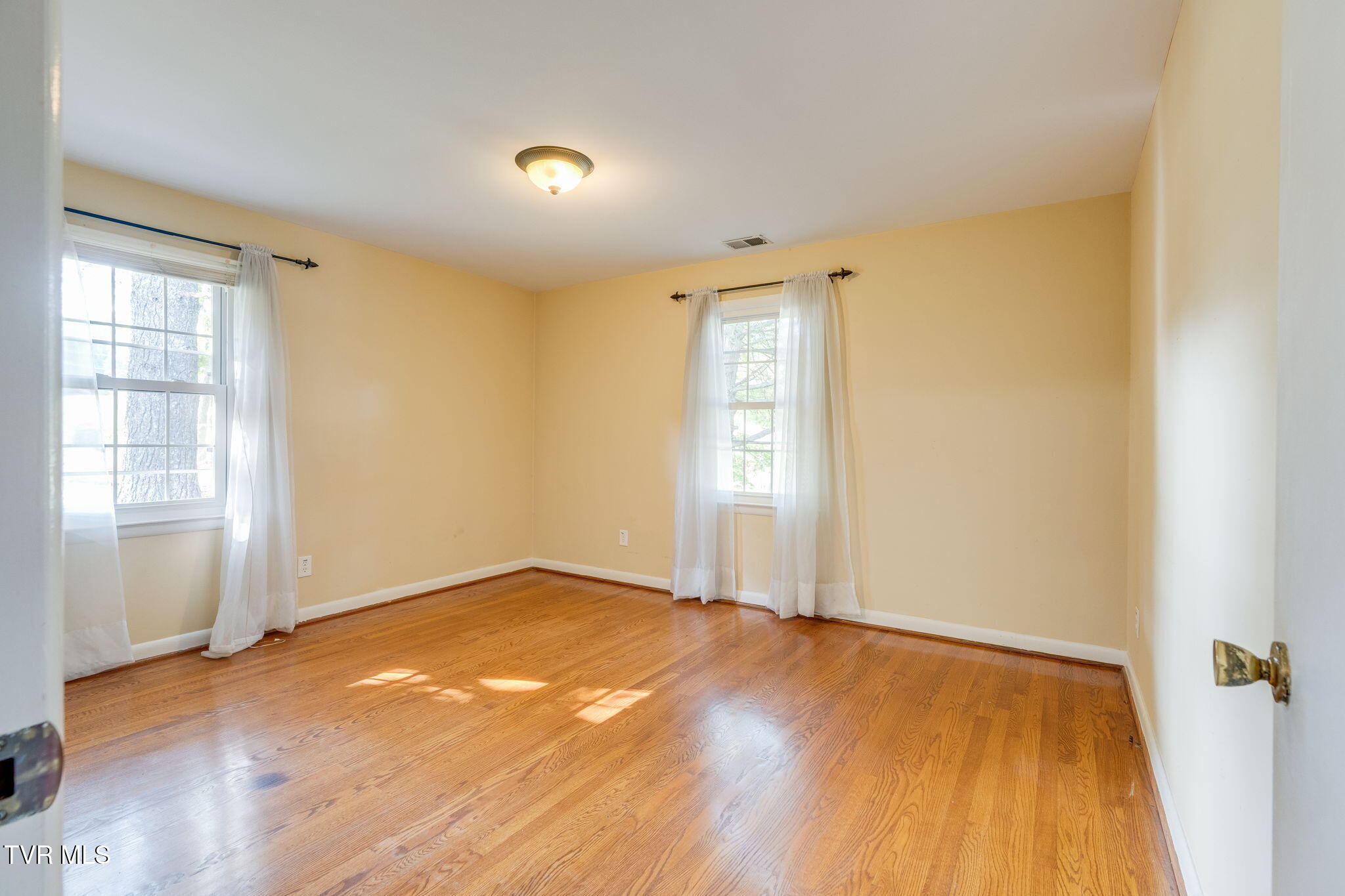 Empty room with hardwood floors, pale yellow walls, and two curtained windows.