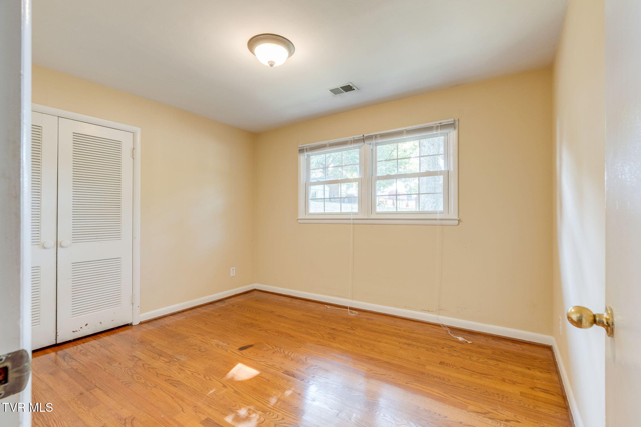 Empty bedroom with hardwood floors, a louvered closet, and two windows.