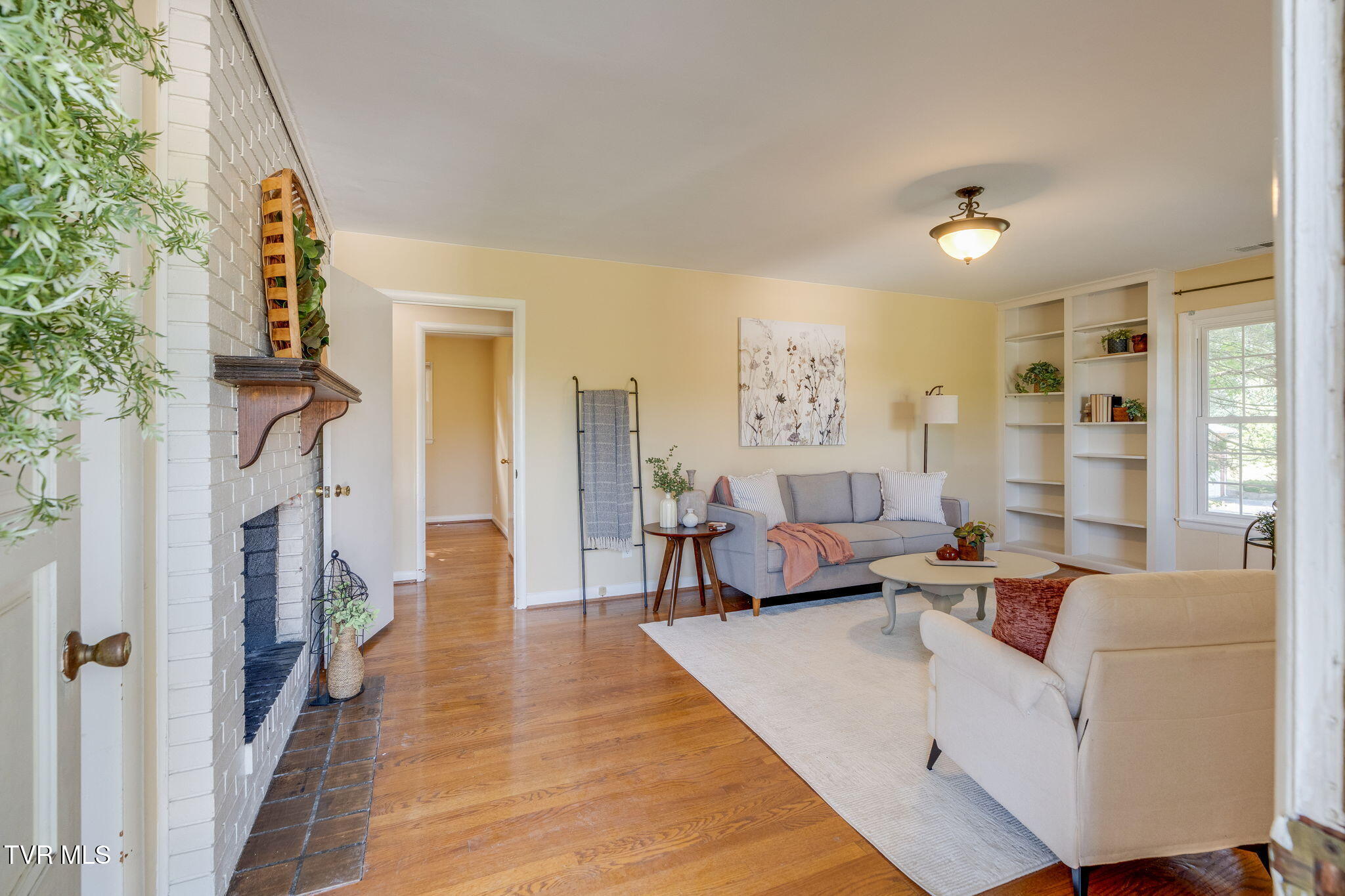 Staged living room with a white brick fireplace, hardwood floors, and built-in shelving.