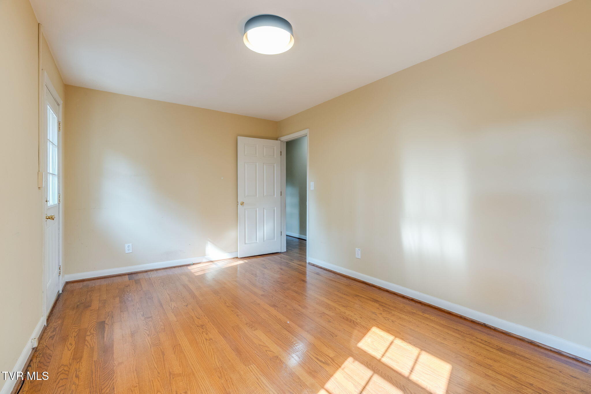 Empty room with hardwood floors, beige walls, and sunlight through a glass-paneled door.
