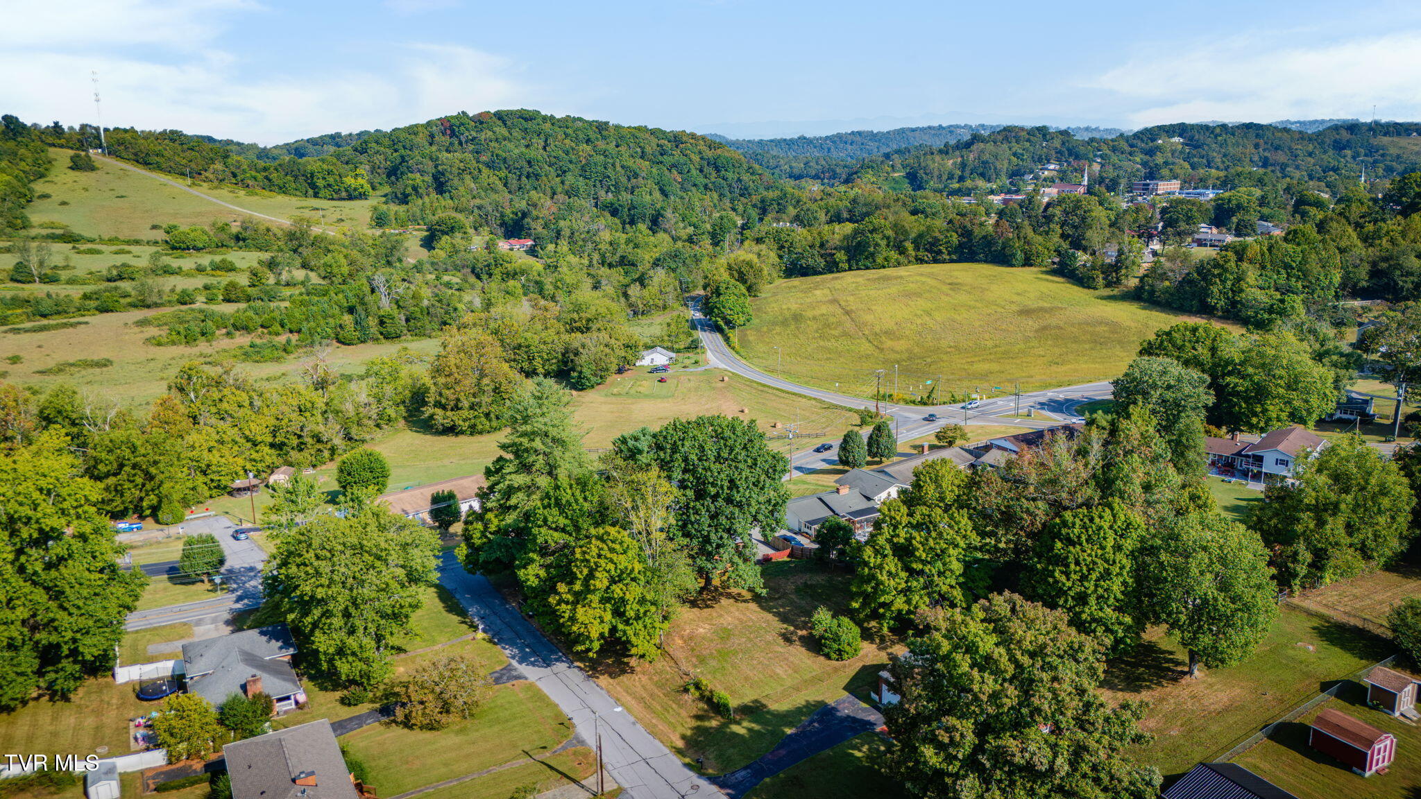 Aerial view of an open field beside wooded hills, homes, and intersecting roads.