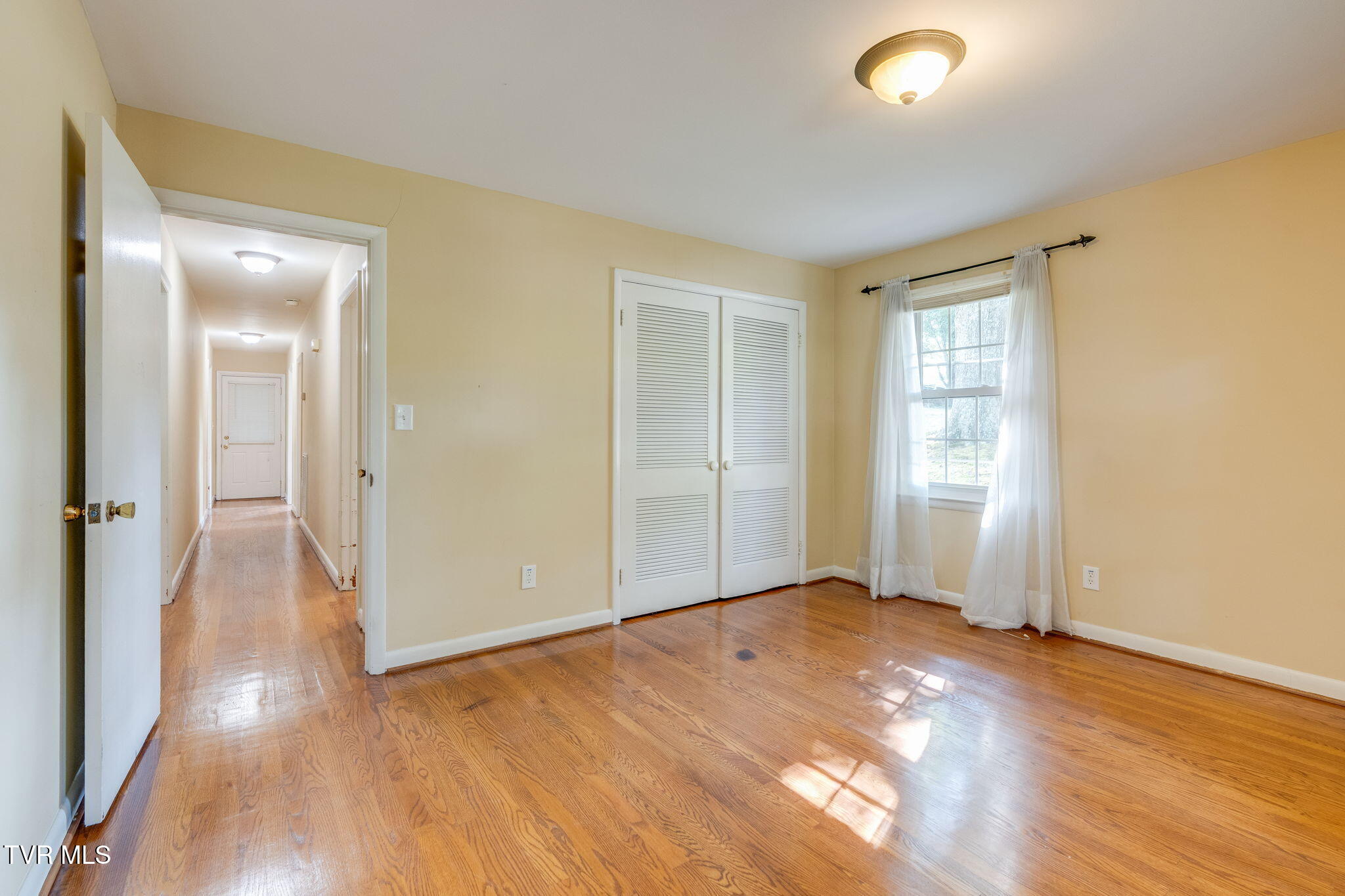 Empty bedroom with hardwood floors, a louvered closet, and a curtained window.