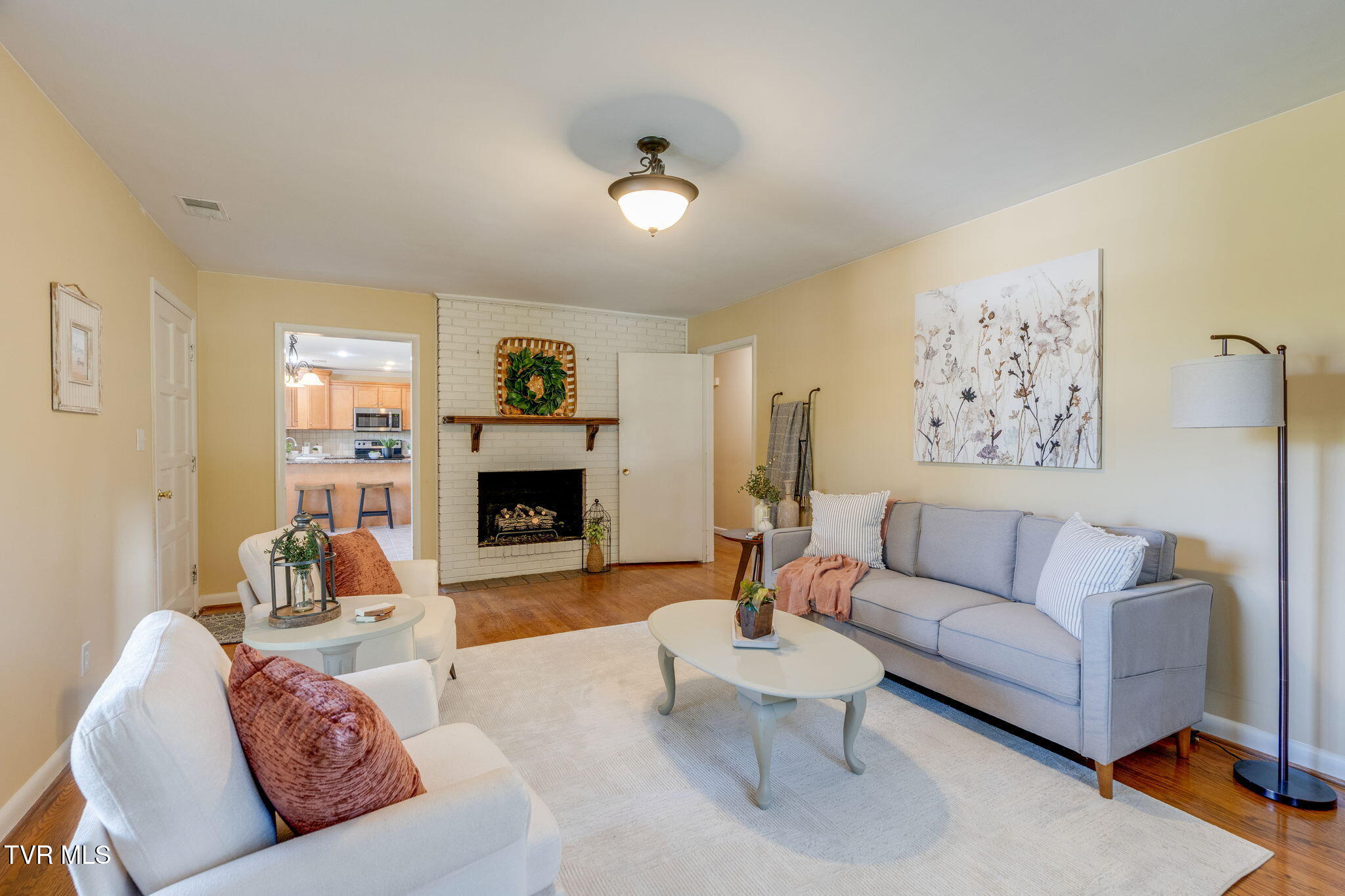 Staged living room with a white brick fireplace, gray sofa, and kitchen visible through the doorway.