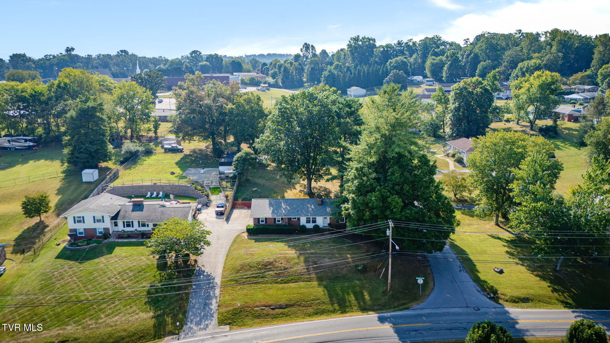 Aerial view of homes and wooded residential lots, with a fenced pool and road in the foreground.
