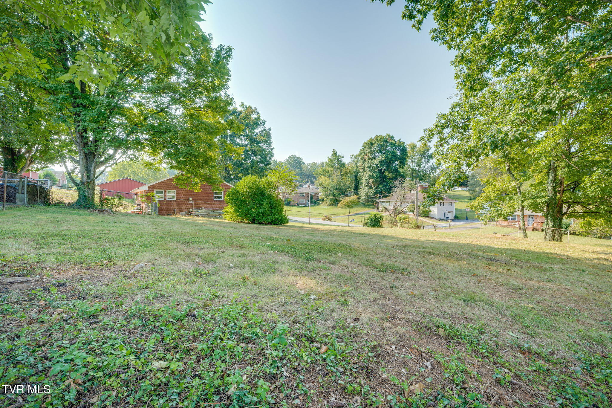 Sloping grassy lot beneath mature trees, with neighboring homes beyond a fence.
